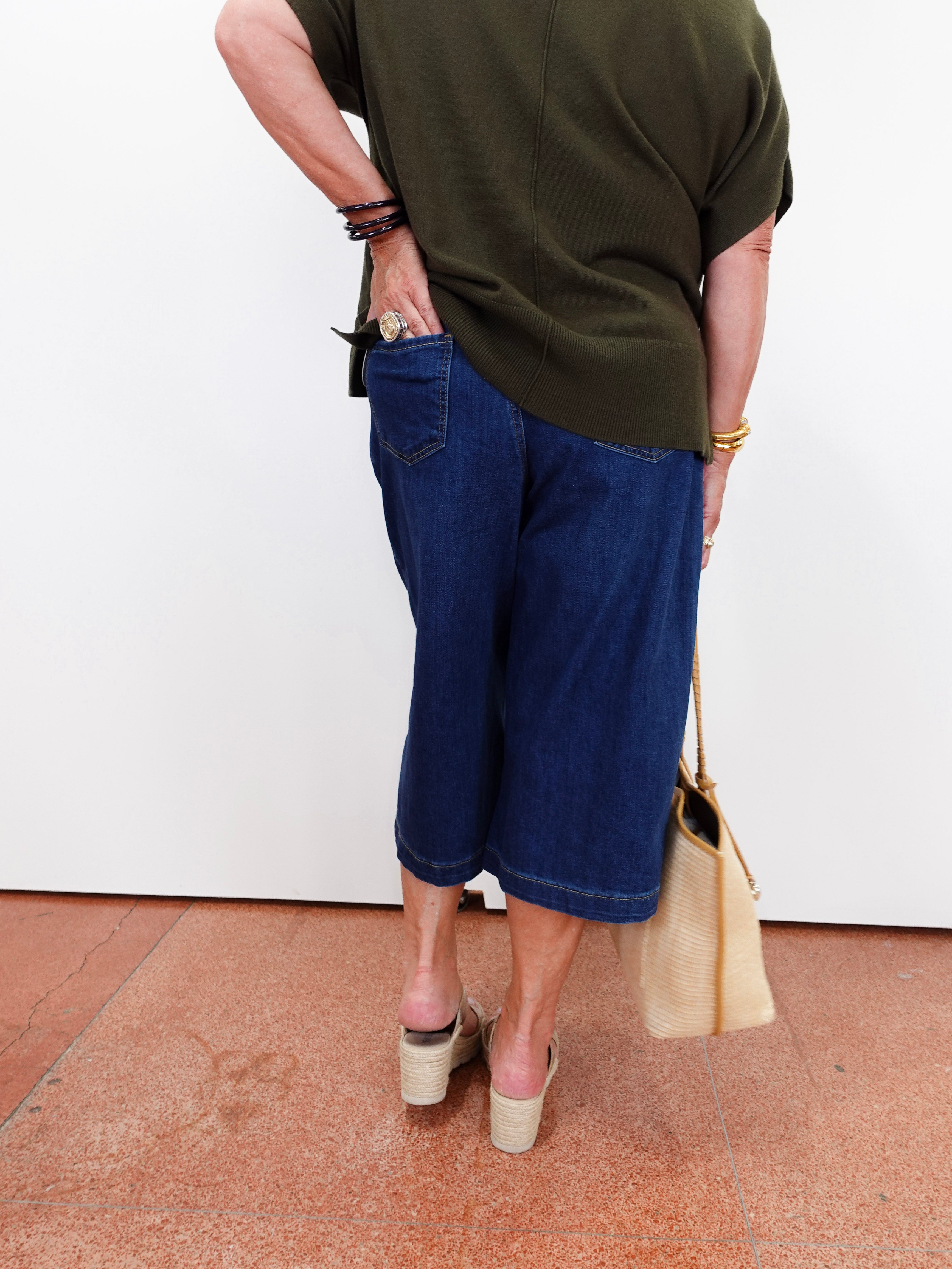 A person shown from the back wearing dark blue cropped denim shorts, an olive green top, and beige wedge sandals, holding a beige tote bag against a white wall.