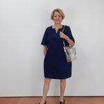 A woman with short blonde hair wearing a navy blue short-sleeve dress and black sandals, holding a clear plastic shoulder bag with a black strap, standing against a plain white wall.