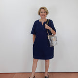 A woman with short blonde hair wearing a navy blue short-sleeve dress and black sandals, holding a clear plastic shoulder bag with a black strap, standing against a plain white wall.