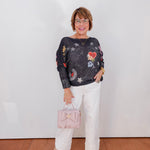 Woman wearing a dark sweater with colorful patterns and white pants, holding a pink handbag against a white wall.