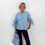 Woman wearing a blue polka dot blouse and black pants, holding a matching handbag against a white background.