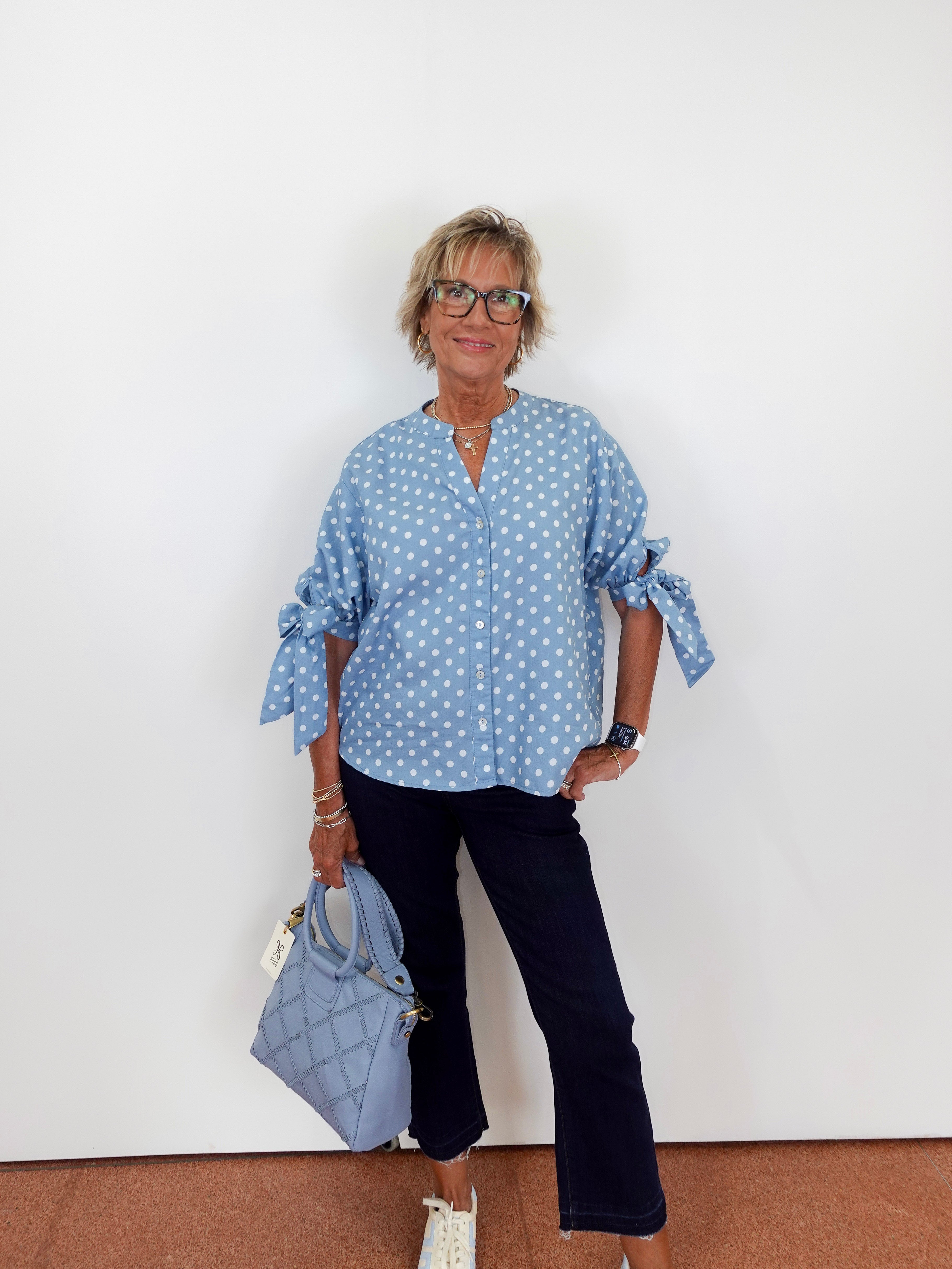 Woman wearing a blue polka dot blouse and black pants, holding a matching handbag against a white background.