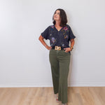 Woman wearing a floral blouse and green pants against a white wall.