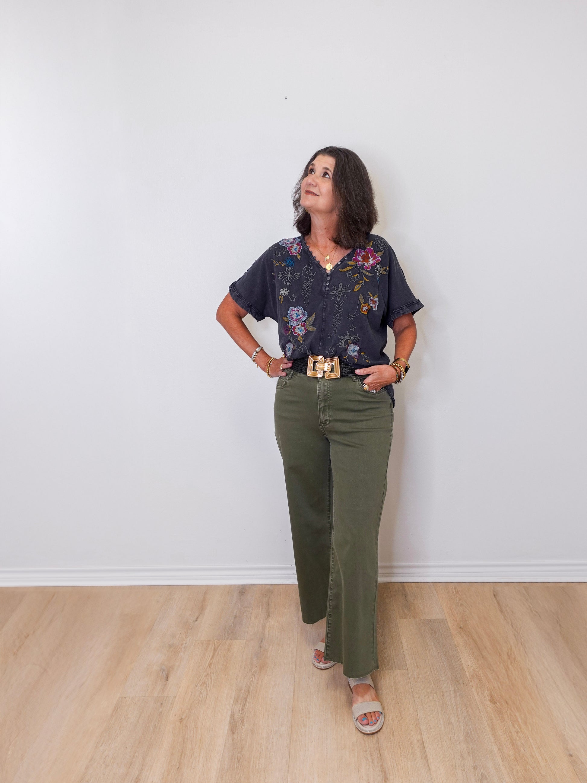 Woman wearing a floral blouse and green pants against a white wall.