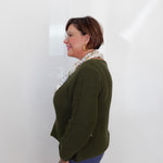 A woman with short brown hair and glasses, wearing a textured olive green cardigan, a patterned cream scarf, and blue pants, standing in profile against a white wall.