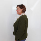 A woman with short brown hair and glasses, wearing a textured olive green cardigan, a patterned cream scarf, and blue pants, standing in profile against a white wall.
