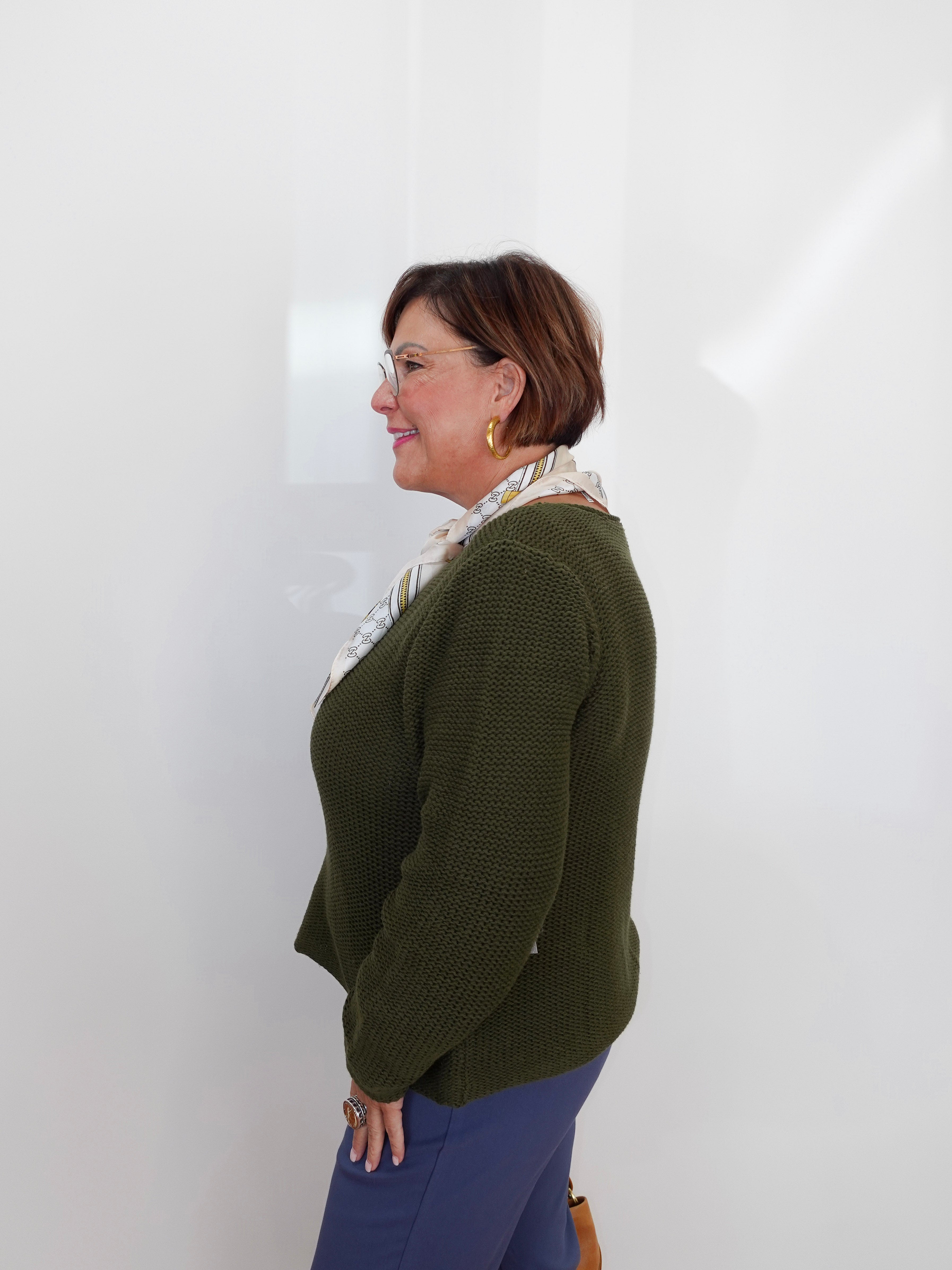 A woman with short brown hair and glasses, wearing a textured olive green cardigan, a patterned cream scarf, and blue pants, standing in profile against a white wall.