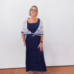 Woman wearing a blue dress with a patterned shawl against a white wall.