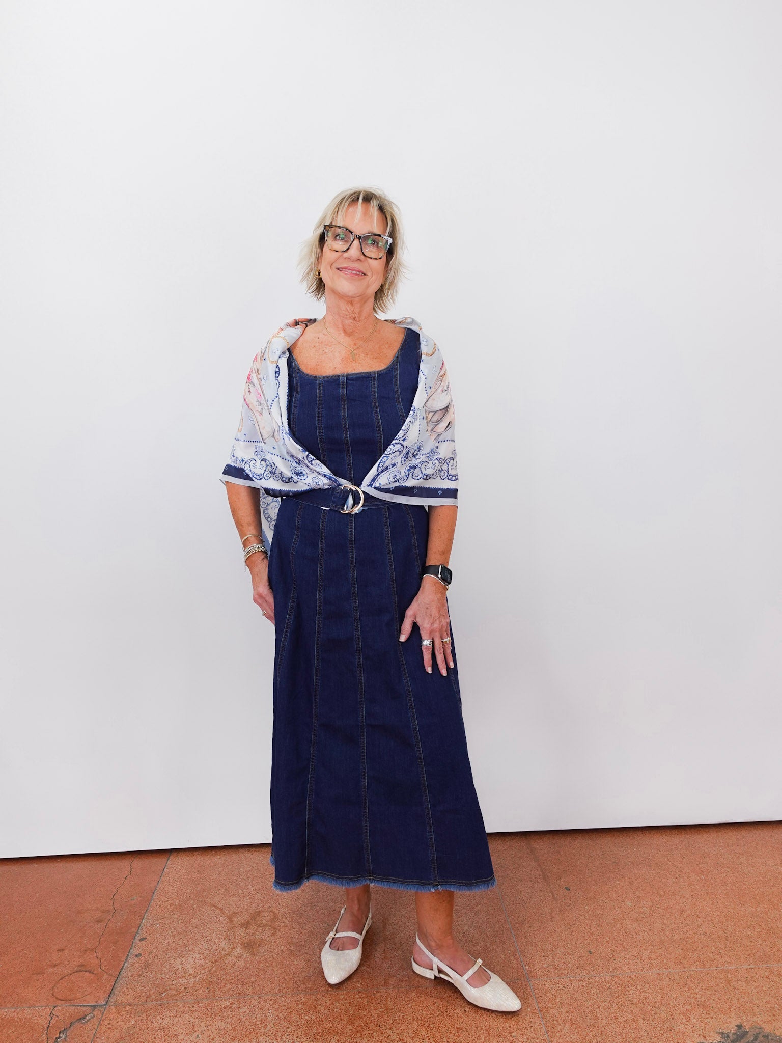 Woman wearing a blue dress with a patterned shawl against a white wall.