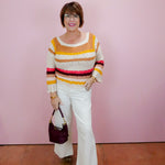 Woman wearing a colorful striped sweater and white pants against a pink wall.