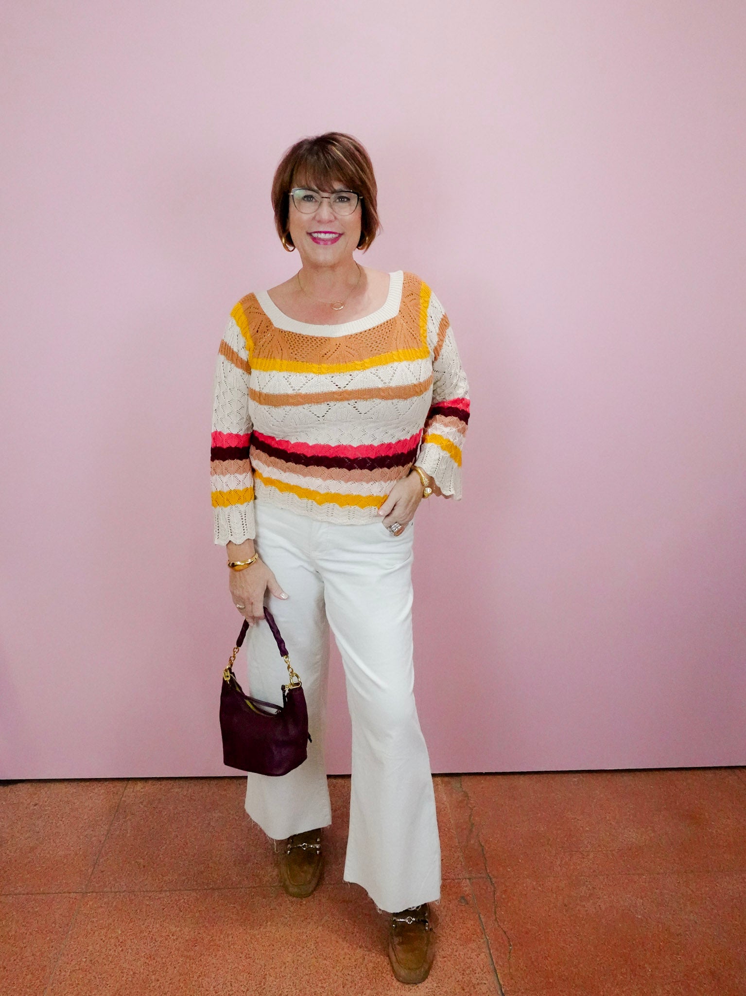 Woman wearing a colorful striped sweater and white pants against a pink wall.
