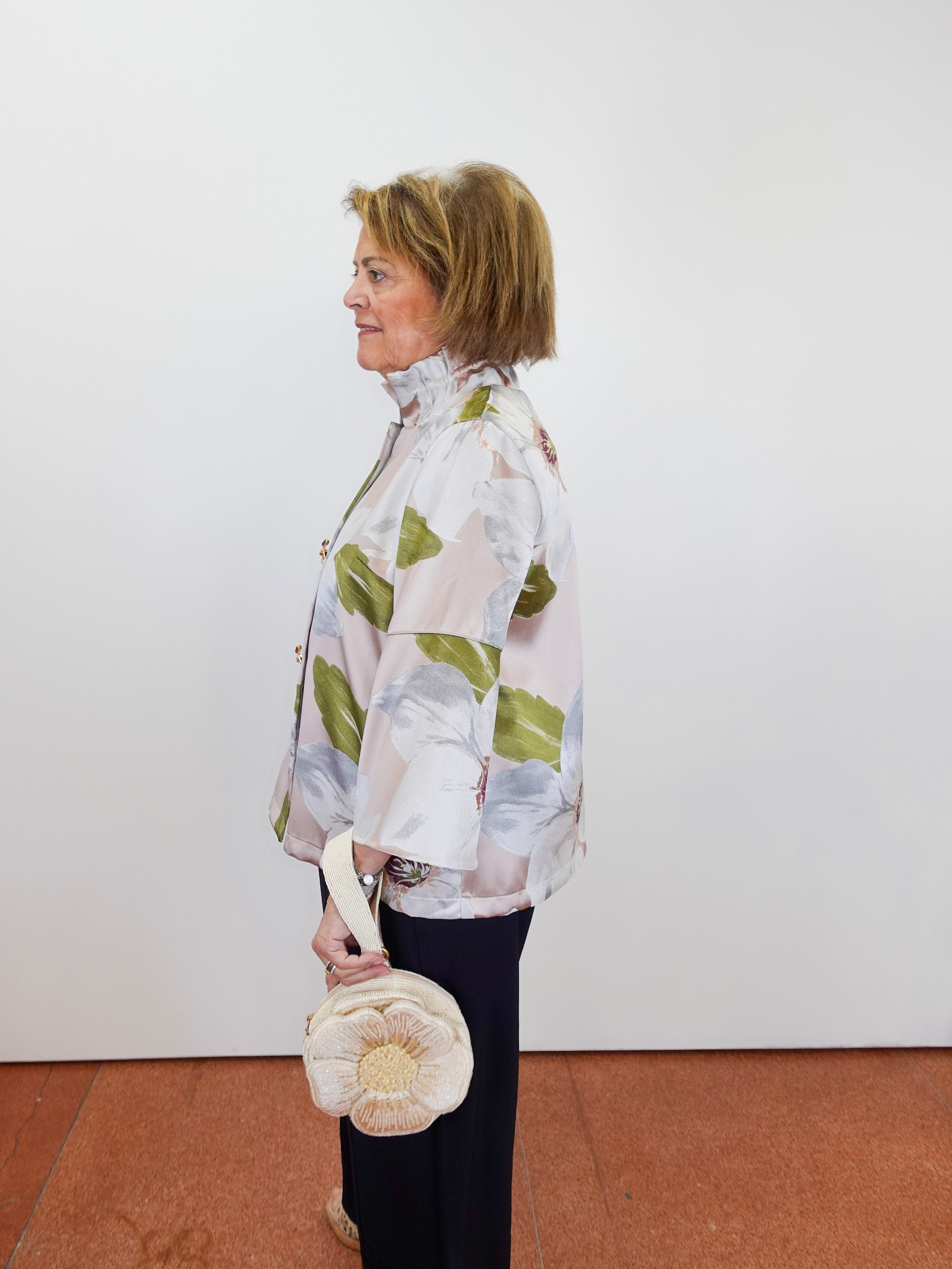 Woman wearing a patterned jacket holding a round bag against a white wall.