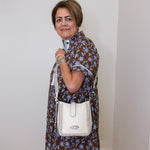 Woman in a floral dress holding a white handbag against a plain wall.