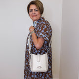 Woman in a floral dress holding a white handbag against a plain wall.