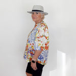 Person wearing a colorful floral shirt and black shorts with a gray hat against a white wall.