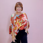 Woman wearing a colorful floral blouse and black pants against a pink background