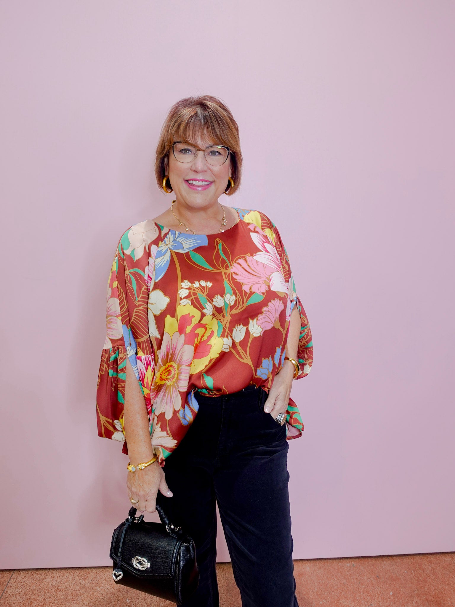 Woman wearing a colorful floral blouse and black pants against a pink background