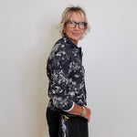 Woman wearing a black and white floral shirt and black pants against a plain white wall.