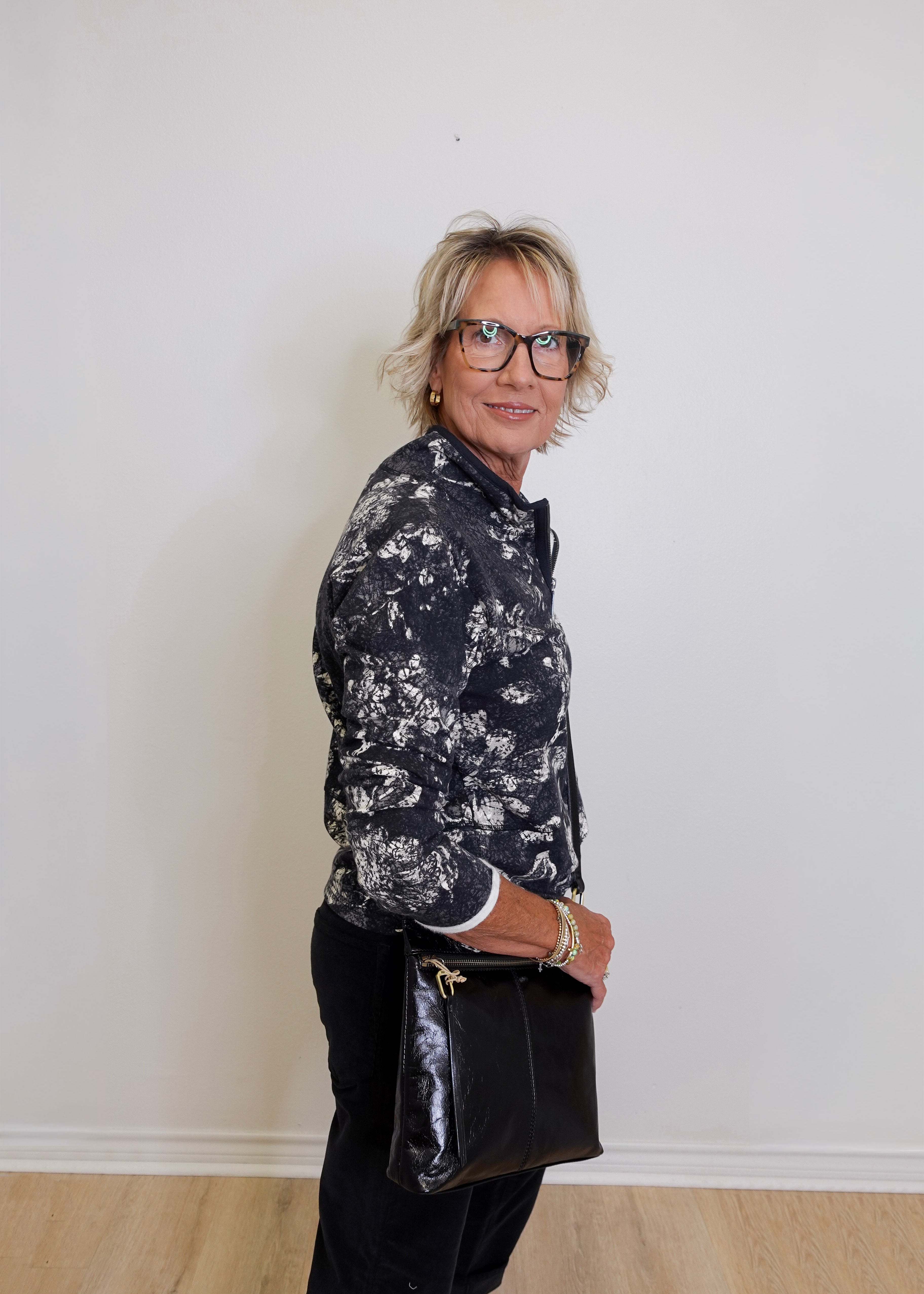 Woman wearing a black and white floral shirt and black pants against a plain white wall.