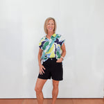 Woman wearing a colorful shirt and black shorts standing against a white wall.