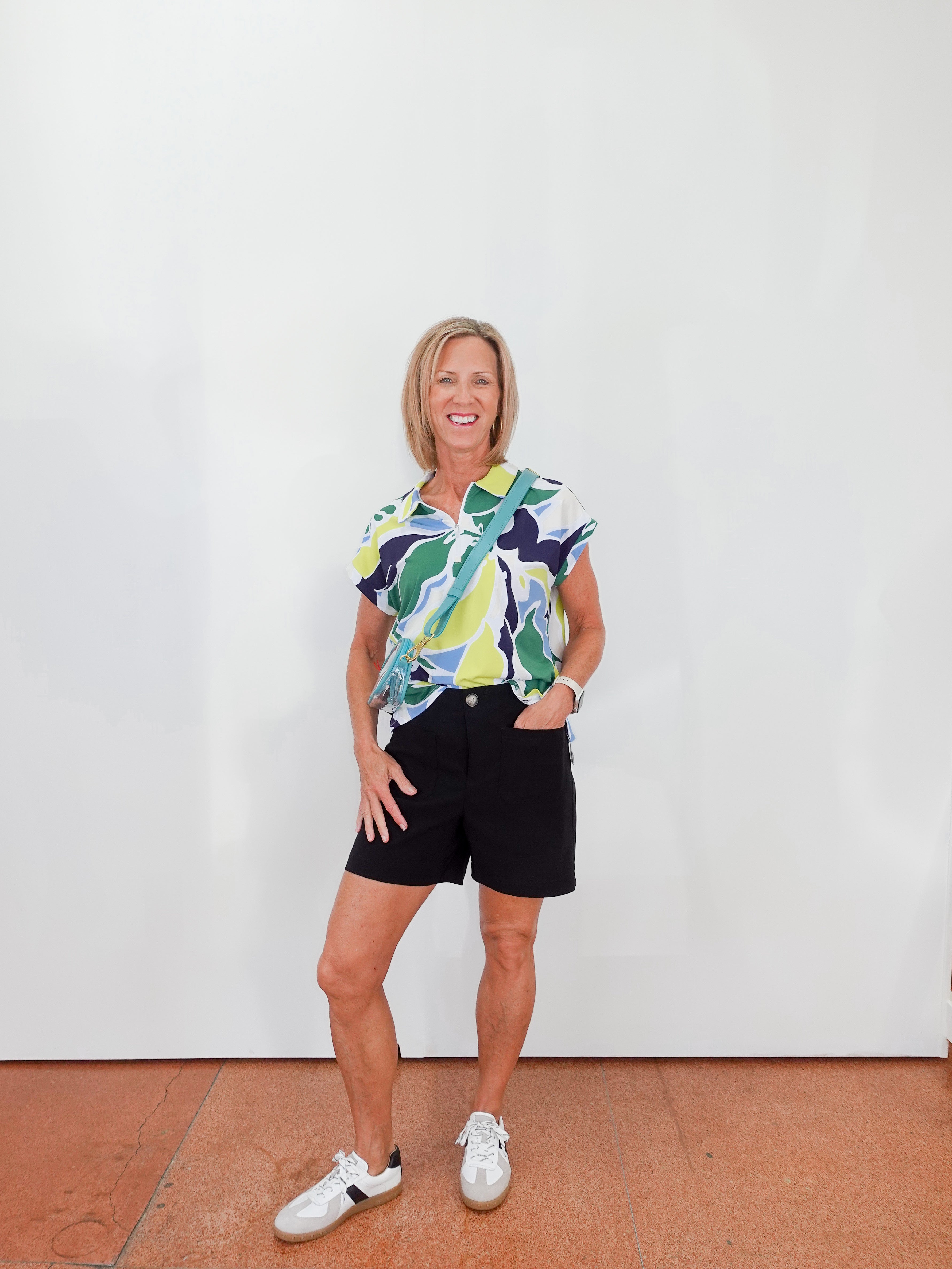 Woman wearing a colorful shirt and black shorts standing against a white wall.