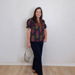 Woman wearing a floral blouse and black pants standing against a white wall.