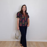 Woman wearing a floral blouse and black pants standing against a white wall.