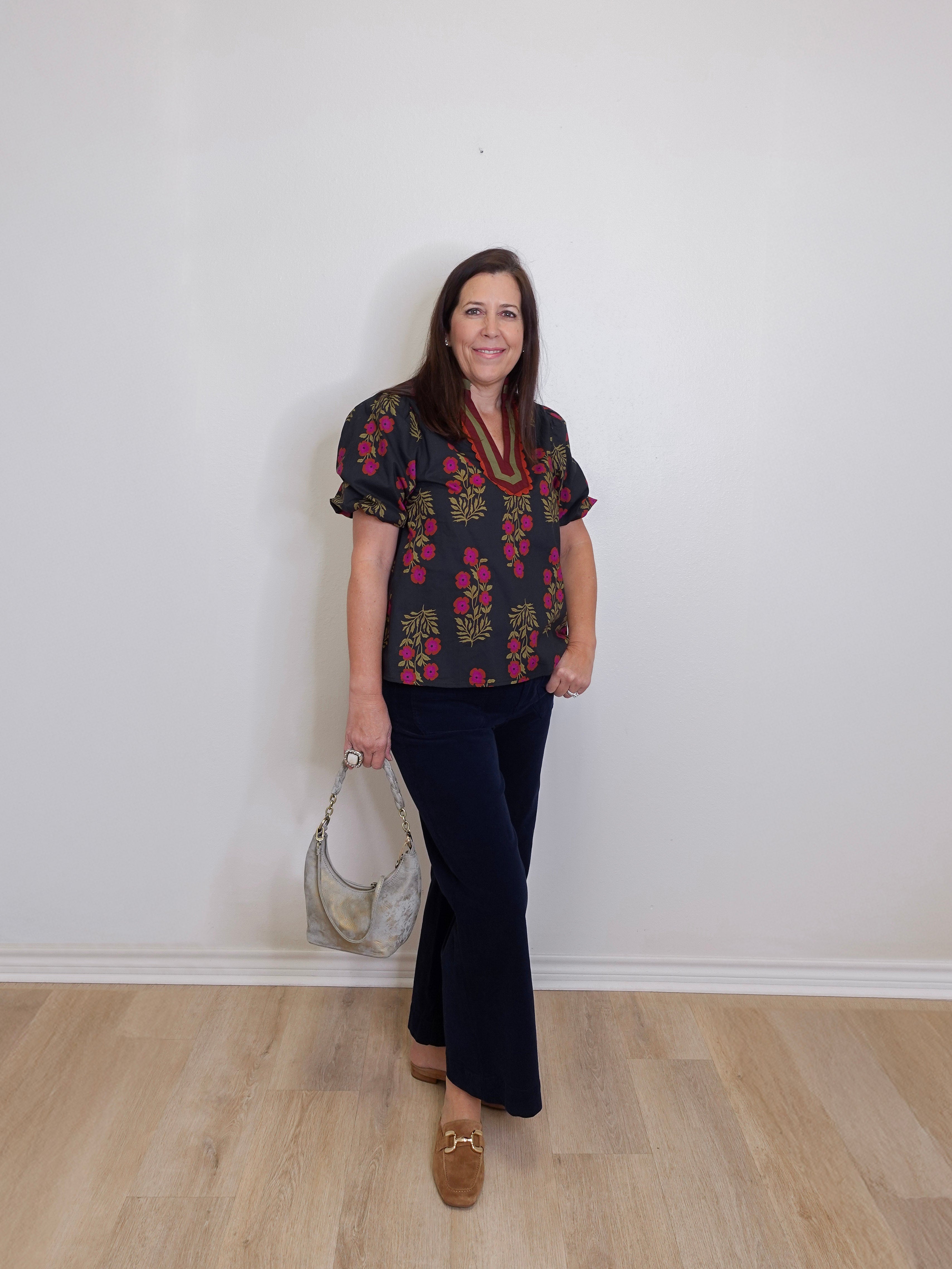 Woman wearing a floral blouse and black pants standing against a white wall.