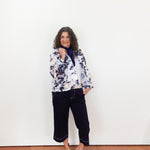 Woman wearing a patterned jacket and black pants against a white wall.