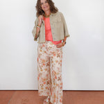 A woman stands against a plain white wall wearing a light beige cropped denim jacket over a coral pink top and cream floral wide-leg pants, paired with beige sandals.