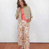 A woman stands against a plain white wall wearing a light beige cropped denim jacket over a coral pink top and cream floral wide-leg pants, paired with beige sandals.