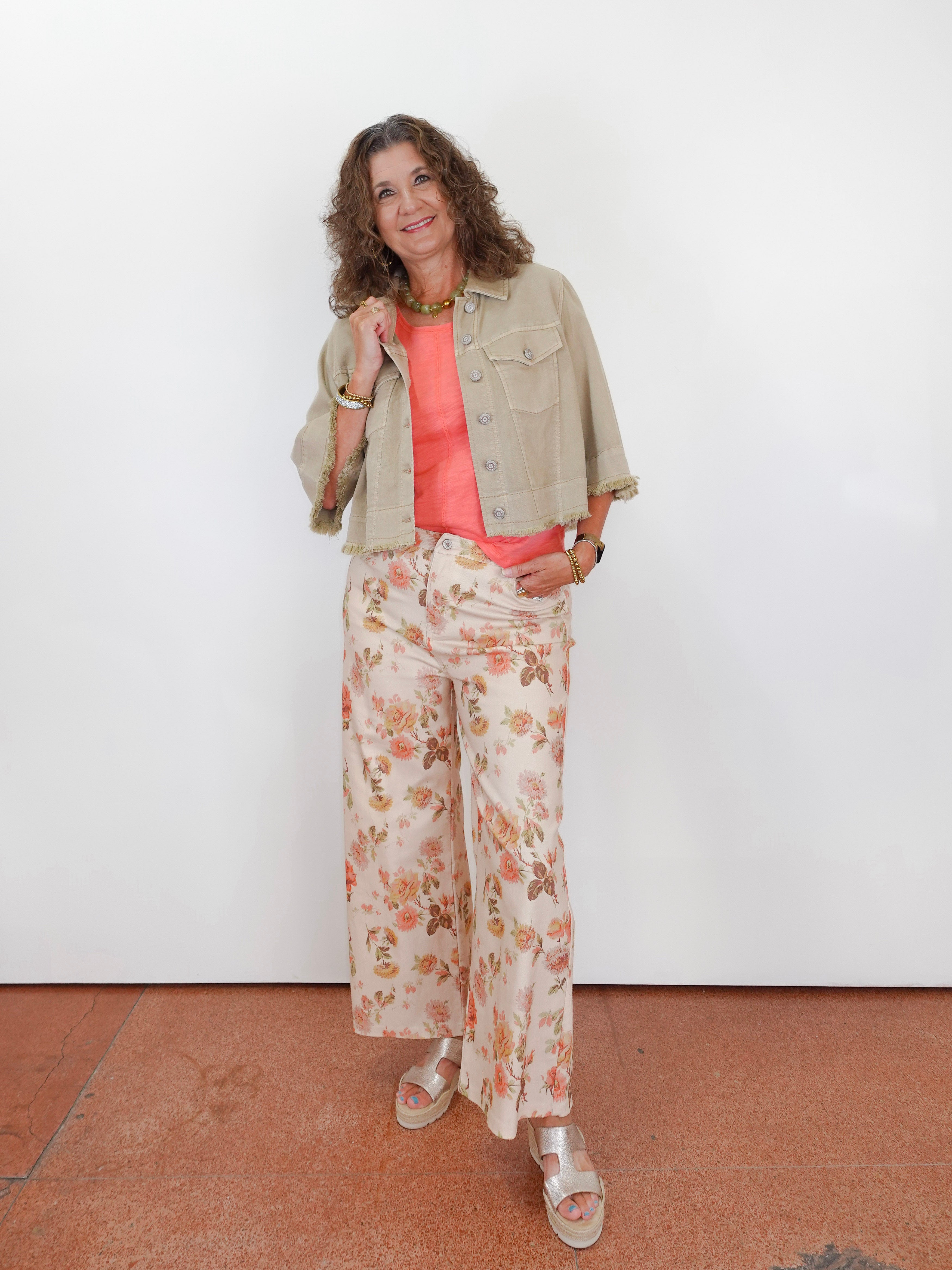 A woman stands against a plain white wall wearing a light beige cropped denim jacket over a coral pink top and cream floral wide-leg pants, paired with beige sandals.