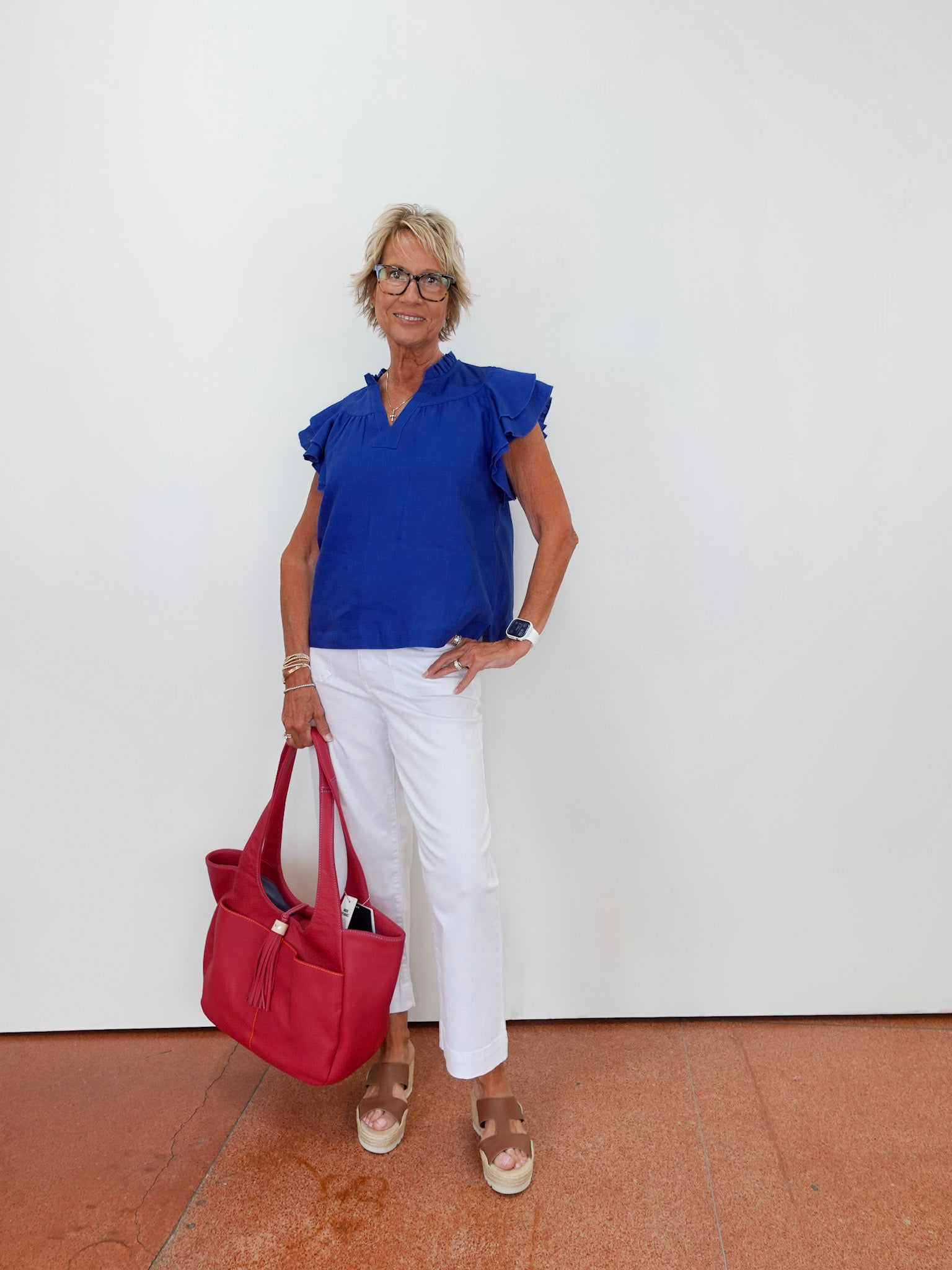 Woman wearing a blue top and white pants, holding a red bag against a white wall.