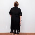 Person wearing a black dress with floral embroidery, standing against a white wall.