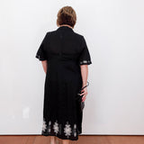 Person wearing a black dress with floral embroidery, standing against a white wall.