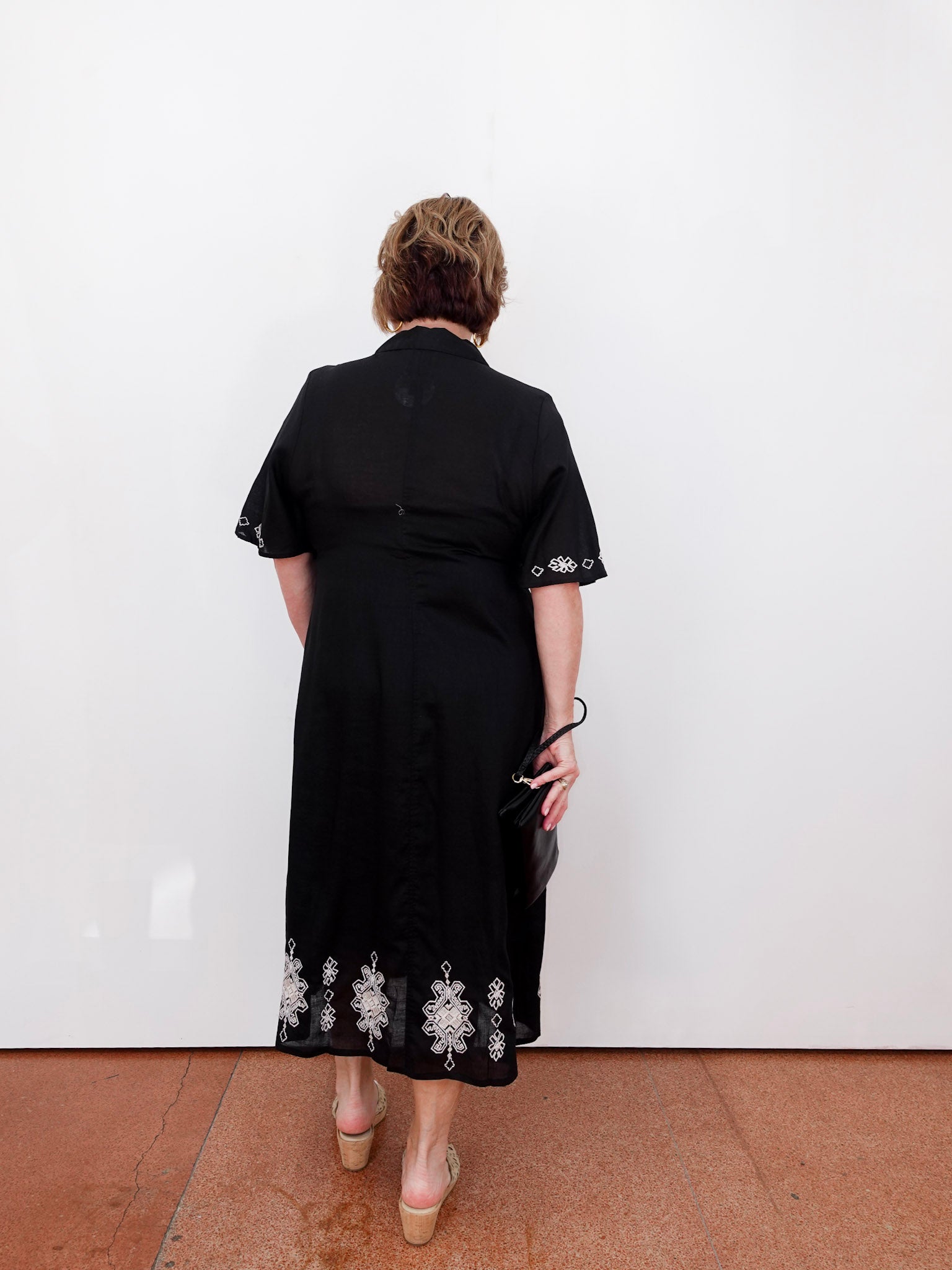 Person wearing a black dress with floral embroidery, standing against a white wall.