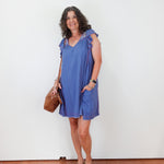 Woman wearing a blue dress holding a brown bag against a white wall.