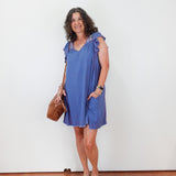 Woman wearing a blue dress holding a brown bag against a white wall.