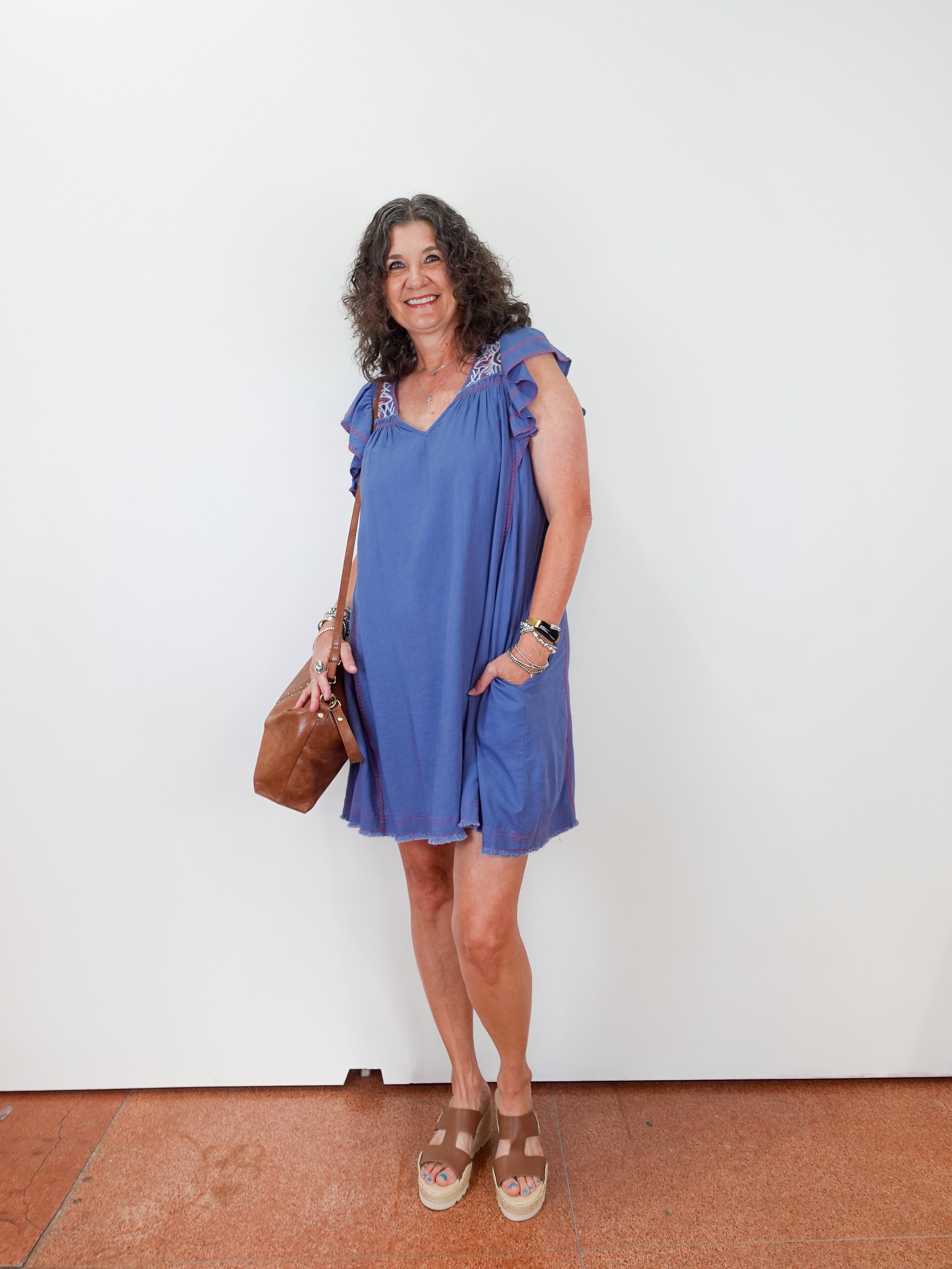 Woman wearing a blue dress holding a brown bag against a white wall.