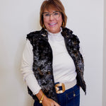 Woman wearing a black floral vest over a white shirt, blue jeans, and glasses against a plain background.