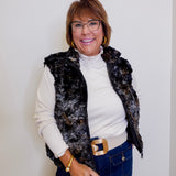 Woman wearing a black floral vest over a white shirt, blue jeans, and glasses against a plain background.
