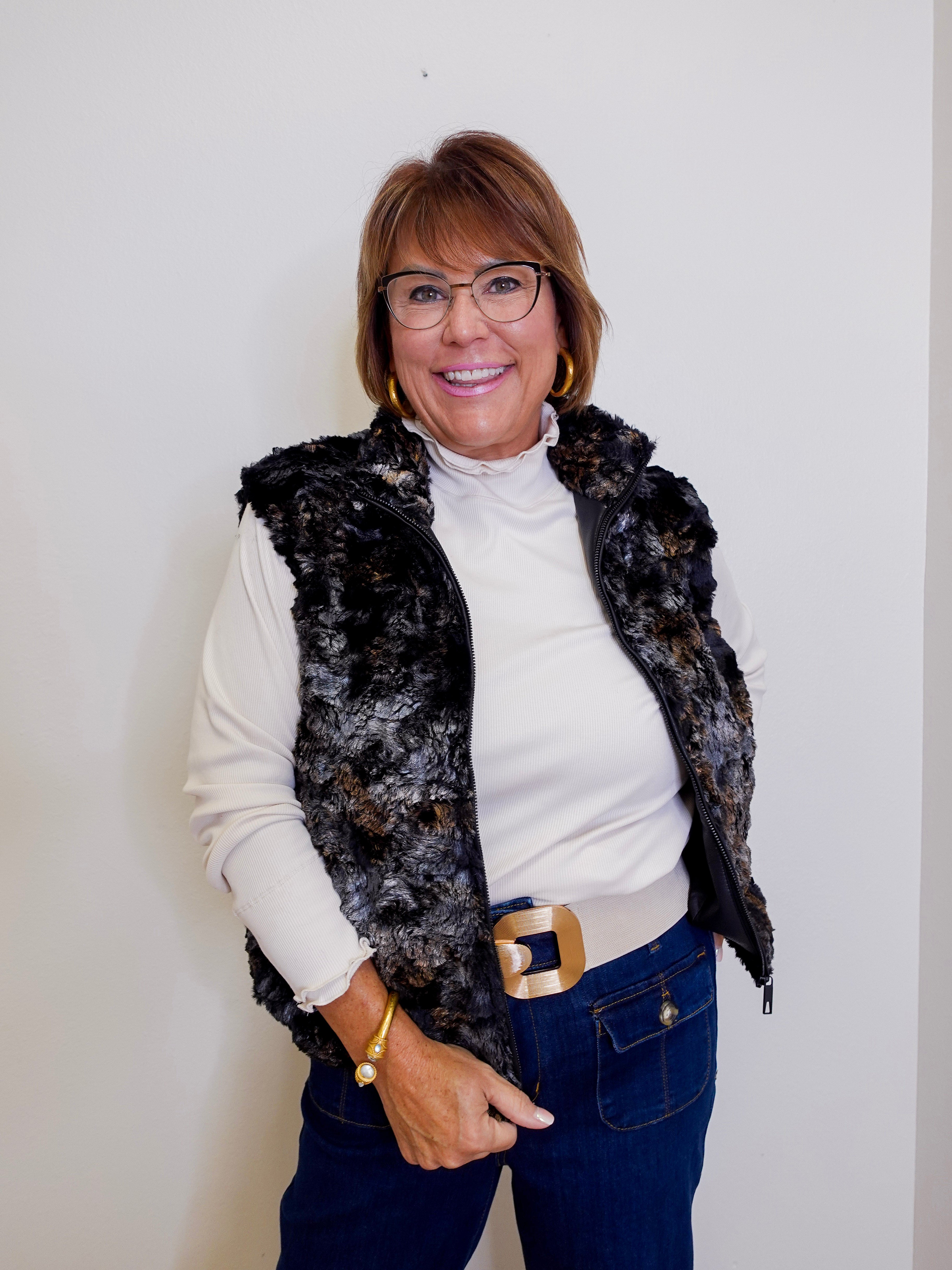 Woman wearing a black floral vest over a white shirt, blue jeans, and glasses against a plain background.
