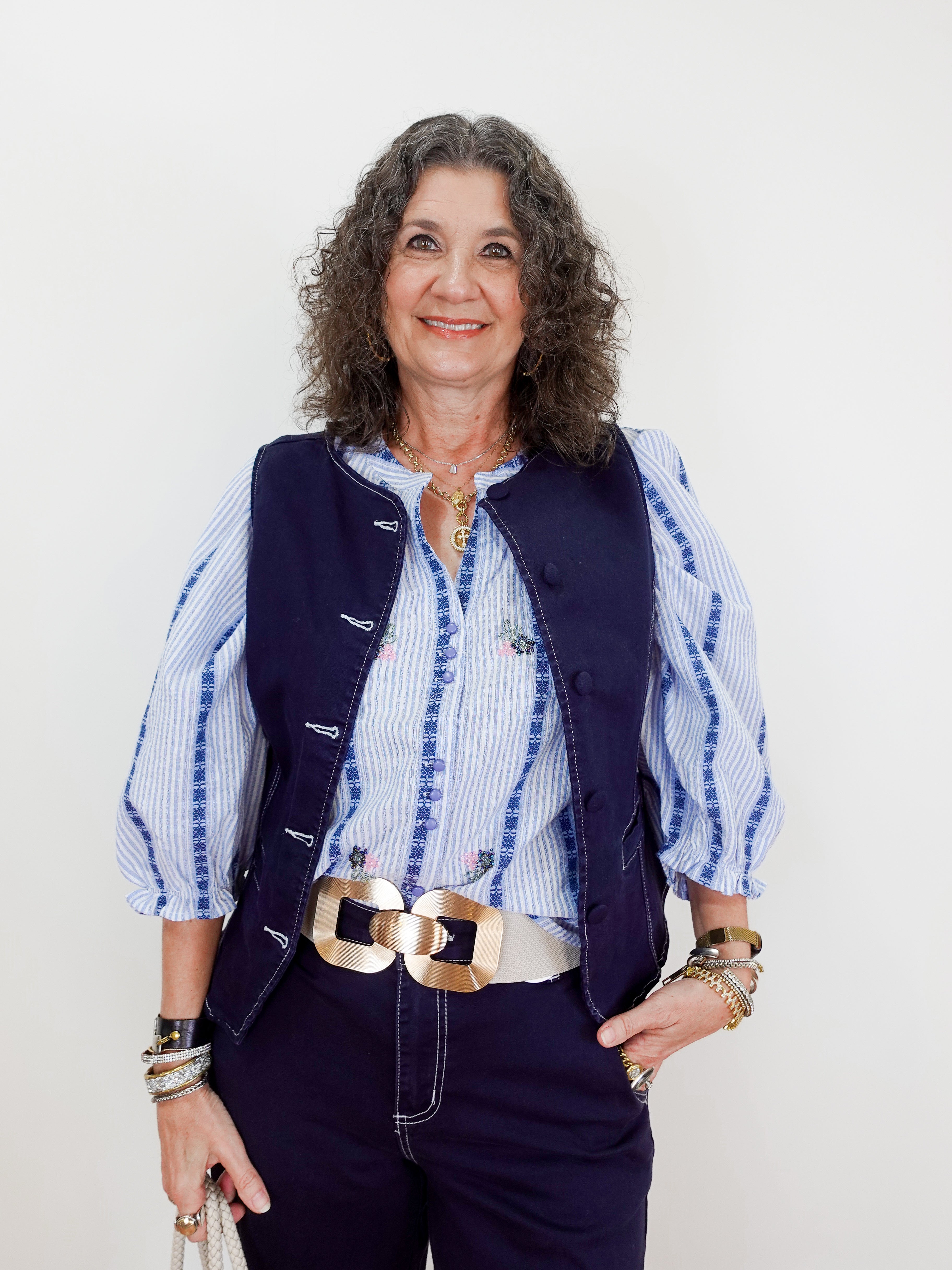 Woman wearing a blue vest over a striped shirt with a gold belt and accessories on a white background