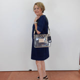 A woman wearing a navy blue dress and black sandals carries a clear tote bag with a leopard print trim and a beige center patch, standing against a plain white wall.