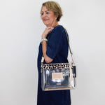 A woman wearing a navy blue dress models a clear rectangular shoulder bag with a leopard-print patterned top band and a silver-toned metal logo plate.