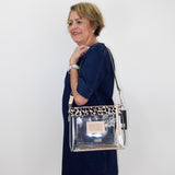 A woman wearing a navy blue dress models a clear rectangular shoulder bag with a leopard-print patterned top band and a silver-toned metal logo plate.