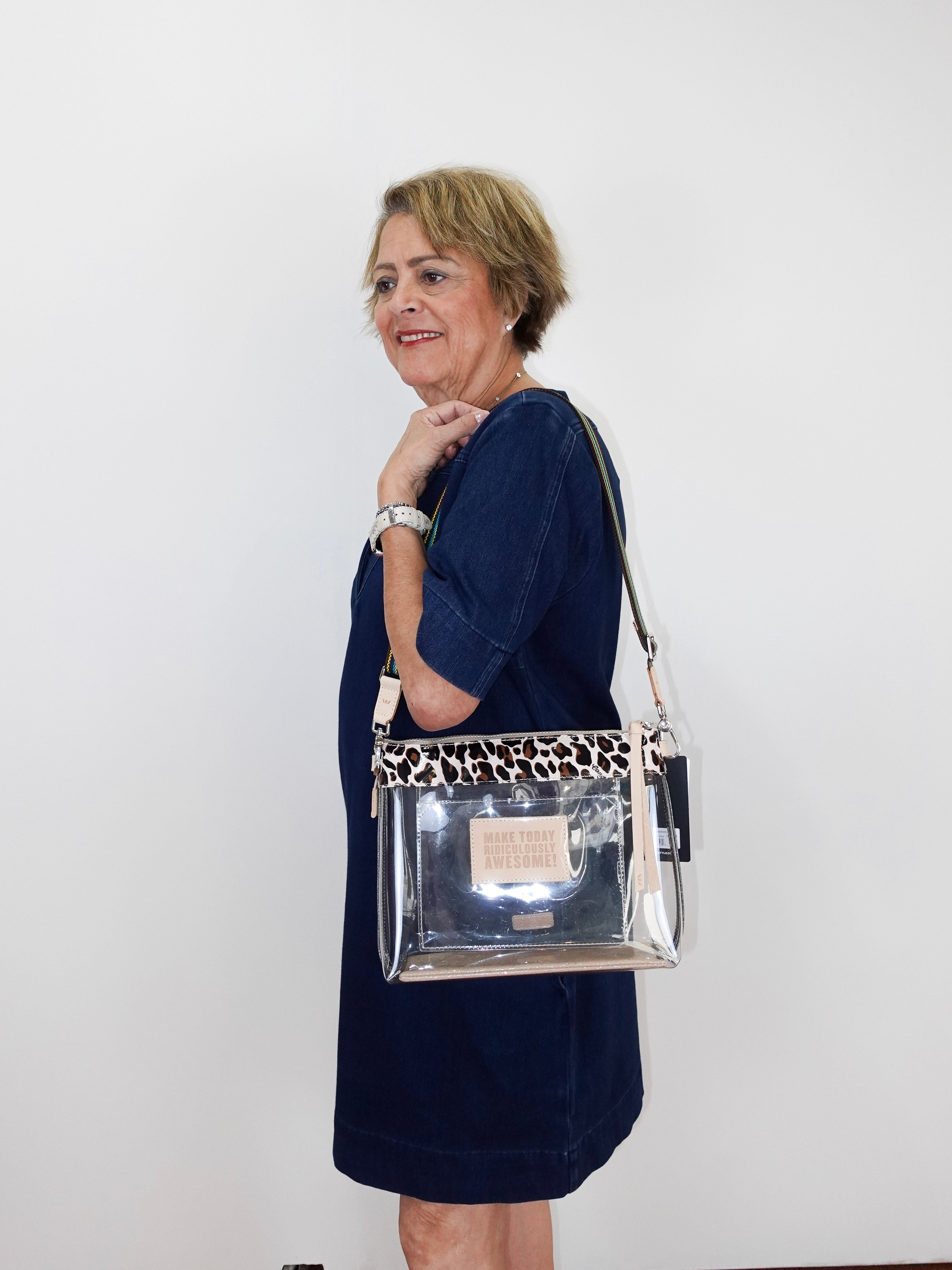 A woman wearing a navy blue dress models a clear rectangular shoulder bag with a leopard-print patterned top band and a silver-toned metal logo plate.