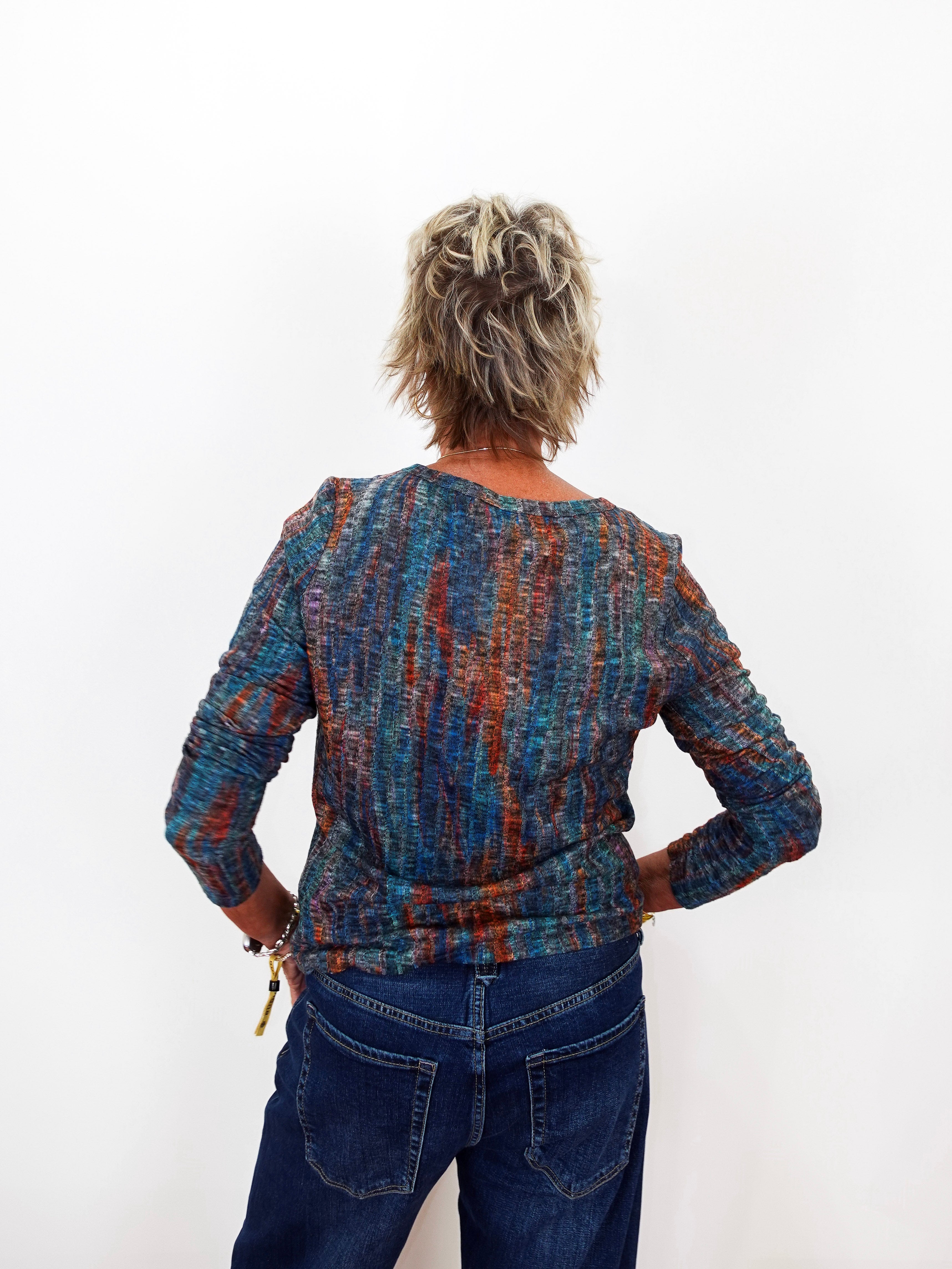A woman viewed from behind wearing a long-sleeve textured knit top with a multicolor abstract pattern in blue, orange, and purple, paired with dark blue jeans against a plain white background.