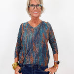 An older woman with short blonde hair and glasses is wearing a long-sleeve V-neck top with a blue, teal, and rust-orange abstract pattern, paired with dark blue jeans. She is posing against a solid white background.