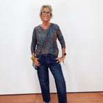 A woman with short gray hair and glasses stands against a white wall wearing a blue and orange patterned long-sleeve top, dark blue jeans, and beige loafers.